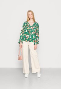 Blusa verde floral com mangas longas, decote em V e punhos franzidos; combinada com calças largas em creme e ténis brancos; segurando uma pequena bolsa rosa.