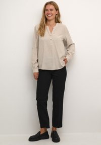Helle beige gestreifte Bluse mit Henley-Ausschnitt, Knopfakzenten und langen Ärmeln, kombiniert mit schwarzen tapered Hosen und schwarzen Slippern.