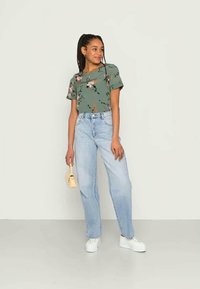 Camisola de manga curta com estampado floral verde, jeans de cintura alta azul claro, ténis brancos, segurando uma pequena bolsa bege. Fundo simples.