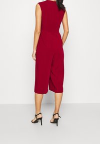Jumpsuit culotte bordeaux avec un design sans manches, fermeture éclair au dos et jambes larges. Associé à des sandales à talons hauts noires. Texture lisse.