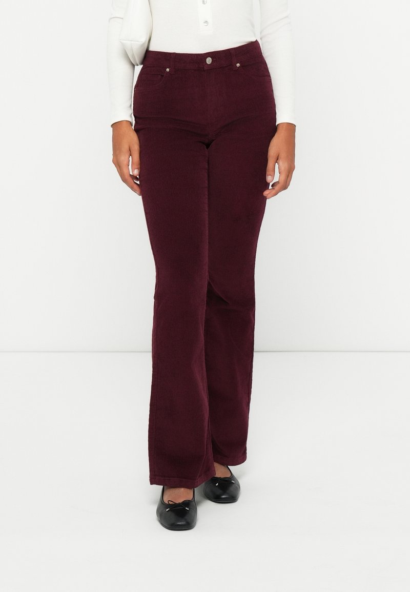Femme portant un pantalon en velours côtelé bordeaux taille haute, associé à des ballerines noires et un haut blanc à manches longues.
