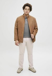 Veste en suédine marron avec fermeture éclair, col montant et manches longues, associée à une chemise grise et un pantalon rose clair. Design texturé et décontracté.