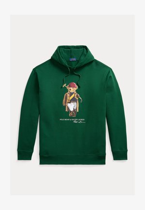 Zöld kapucnis pulóver macival, aki piros sisakot, kabátot és fehér nadrágot visel, valamint egy poloütőt tart a kezében, alatta a Polo Bear by Ralph Lauren felirat.