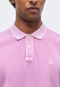 Άνδρας που φοράει ανοιχτό μωβ πόλο πουκάμισο με δύο κουμπιά κουμπωμένα και ένα μικρό λευκό λογότυπο «MOP» στο αριστερό στήθος.