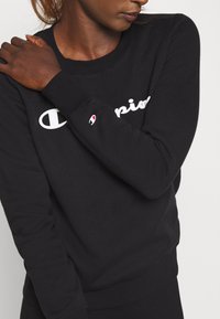 Sudadera negra con cuello redondo, tela suave y puños acanalados. Presenta el logo blanco de "Champion" en el frente y un detalle del logo en la manga.