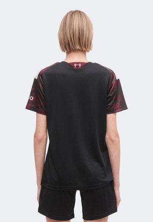 Persona rubia con cabello corto vistiendo pantalones cortos deportivos negros y una camiseta negra y burdeos de manga corta, de espaldas sobre un fondo liso.