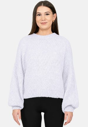 Vrouw met stijl bruin haar, gouden oorringen, een witte dikke gebreide trui met ballonmouwen en zwarte broek, staand met gezicht naar voren.