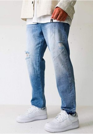 Person iført lyseblå slidte jeans med hvide sneakers og en beige jakke, stående mod en ensfarvet hvid baggrund.