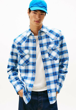 Camicia a quadri blu e bianca con due tasche frontali, bottoni a pressione e yoke in stile western. Abbinata a una t-shirt bianca e pantaloni in denim.