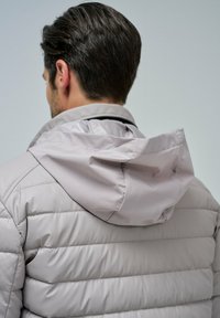 Chaqueta de plumas gris claro con patrón de acolchado horizontal, capucha ajustable y textura suave, que cuenta con un cuello alto y detalle de cremallera.