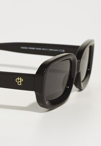 Black, oversized square sunglasses with dark lenses, glossy plastic frame, and yellow logo detail on the temple.