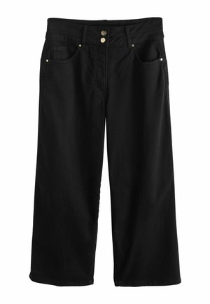 Moteris žingsniuoja pirmyn, vilkėdama berankovį baltą aukštu kaklu modelį, plačias juodas aukšto liemens kelnes, juodus sandalų ir auksinį apyrankę.