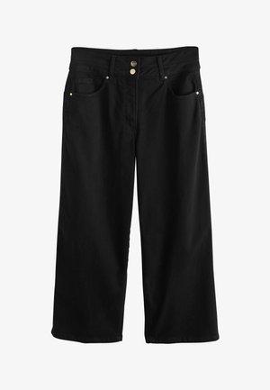 Mujer caminando hacia adelante con top blanco sin mangas y cuello simulado, pantalones negros de pierna ancha y cintura alta, sandalias negras y pulsera dorada.