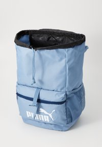 Puma backpack in light blue fabric with a drawstring closure, front zippered pocket, and mesh side pocket. Features logo prominently displayed.