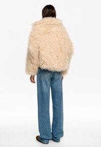 Chaqueta de piel sintética beige con textura de pelaje desaliñado, forma oversized y hombros caídos, combinada con vaqueros de denim azul de pierna ancha.