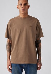 Bruine katoenen t-shirt met ronde hals, korte mouwen en een subtiel geborduurd logo op de borst. Geweven stof met een ontspannen pasvorm.
