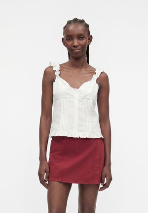 Jeune femme aux cheveux tressés portant un haut blanc sans manches à volants et une mini-jupe rouge, debout devant un fond uni.