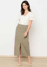 Jupe midi vert olive avec une fente frontale et des passants de ceinture, associée à un chemisier blanc texturé à manches courtes et boutons.