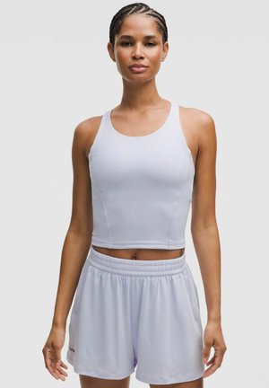 Weißes sportliches Crop-Top mit breiten Trägern, kombiniert mit helllila Shorts mit elastischem Bund und Seitentaschen. Glatte Stofftextur.