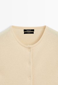 Beige Wollstrickjacke mit Rundhalsausschnitt, fünf Knöpfen und weicher, strukturierter Oberfläche. Markenetikett innen am Kragen sichtbar.