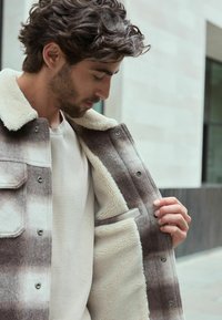 Brown and white plaid wool jacket with shearling lining. Features button closure, chest pockets, and a cozy interior texture.