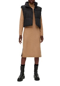 Zwarte puffer vest over een bruine, lange jurk van knie lengte met zijsplitten. Model draagt zwarte enkellaarsjes met dikke zolen.