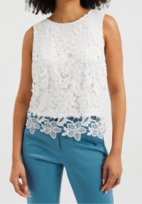 Valkoinen pitsinen hihaton toppi, jossa on kukkakuosi ja aallokkomuotoinen helma, yhdistettynä teal-värisiin housuihin. Raikas, läpikuultava materiaali yksityiskohtaisilla korostuksilla.