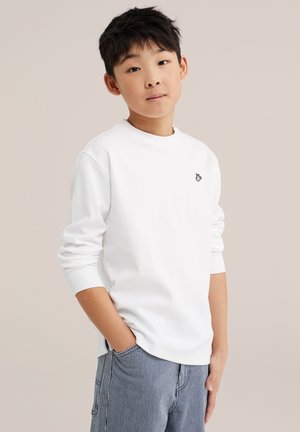 Mladi dječak s kratkom crnom kosom, nosi bijelu košulju dugih rukava i plave hlače na pruge, stoji s jednom rukom u džepu.