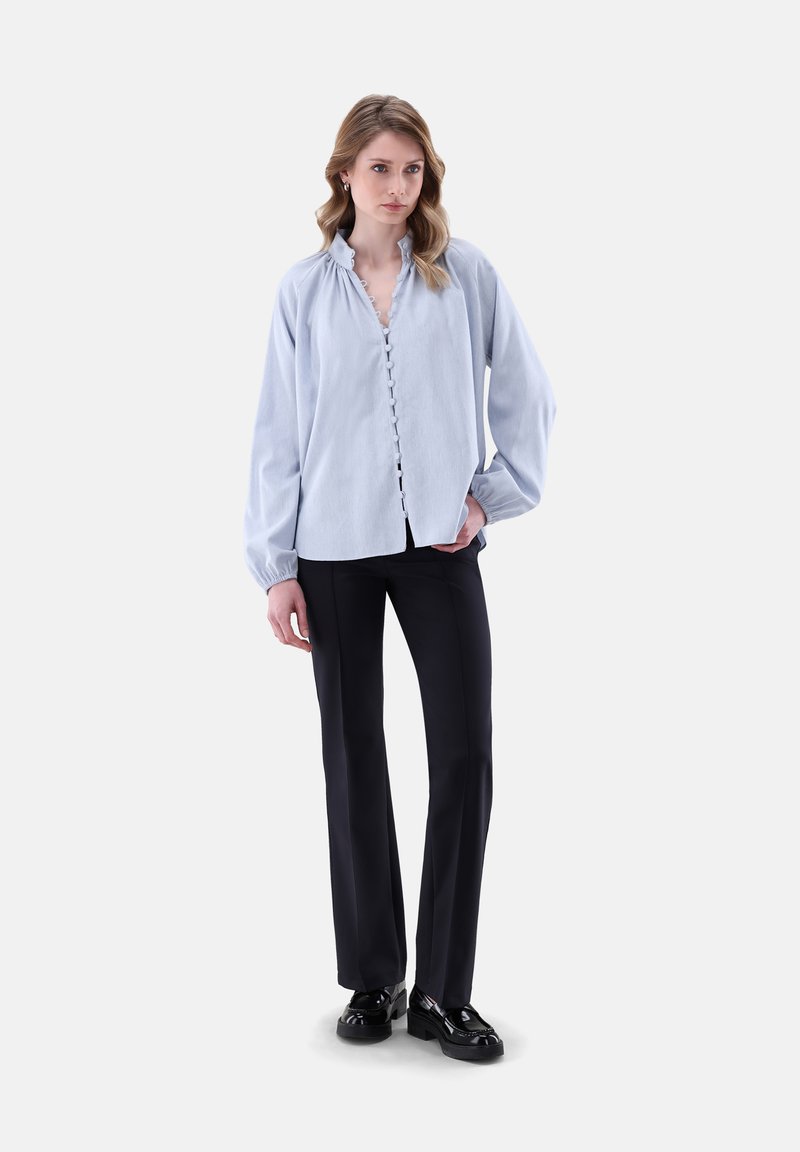 Blusa azzurra chiara con colletto arricciato e maniche ampie, abbinata a pantaloni neri e scarpe nere lucide.