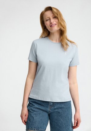 Uśmiechnięta kobieta z rudymi włosami, ubrana w jasnoniebieski krótki rękaw T-shirt i wysokiej talii niebieskie dżinsowe szorty na jednolitym białym tle.