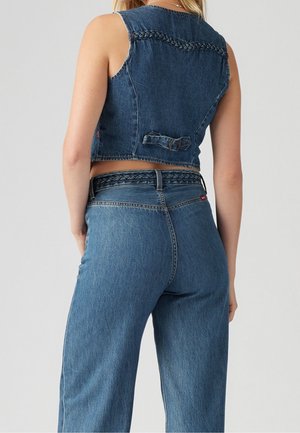 Kvinde iført blå denim ærmeløs kort vest og højttaljede jeans med flettede detaljer på vesten og linningen, stående mod en ensfarvet baggrund.