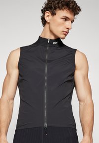 Gilet sans manches noir en tissu léger et résistant à l'eau, avec un col haut et un zip à l'avant, portant le logo "poc" au niveau du col.