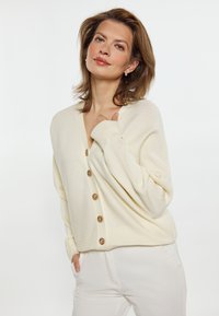 Crème-kleurige, geribde cardigan met een V-hals, houten knopen en losse mouwen. Gecombineerd met lichtgekleurde broek.