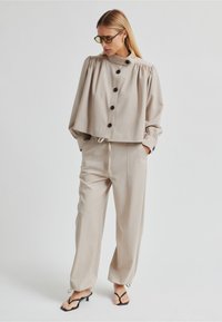 Béžová krátká halenka s nafouklými rukávy a černými knoflíky, doplněná o světle béžové kalhoty se stahovací šňůrkou. Modelka má na sobě černé sandály na podpatku.