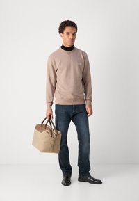 Beige sweatshirt med rund hals, mørkeblå jeans og sorte sko. Holder en tan og brun duffelbag med læderdetaljer.