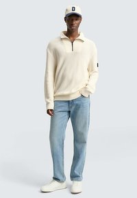 Pulover texturat de culoare crem, cu guler cu fermoar pe jumătate, asortat cu blugi deschis la culoare și adidași albi. Șapca casual completează ținuta.