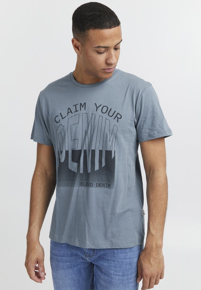 Lyseblå bomullstrøye med korte ermer, med en fremtredende grafikk som leser "CLAIM YOUR DENIM" i mørkegrå og svarte aksenter.