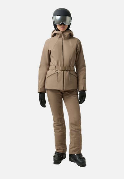 Beige Skijacke mit einem Gürtel, Reißverschlusstaschen und einer Kapuze, kombiniert mit passenden Hosen sowie schwarzen Skistiefeln und Handschuhen und einem schwarzen Helm.