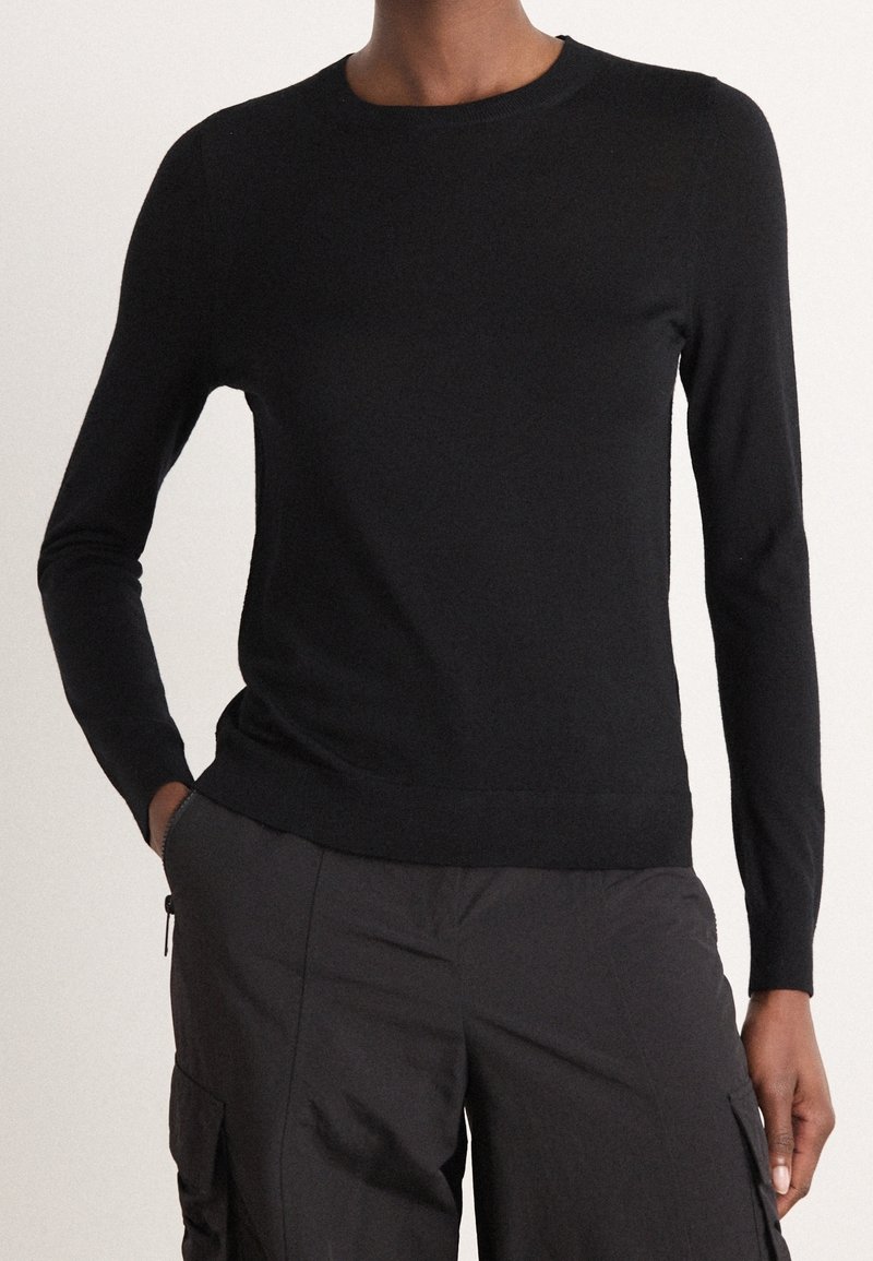 Personne portant un haut noir ajusté à manches longues et col rond, ainsi qu'un pantalon cargo foncé, les mains dans les poches, sur un fond uni.