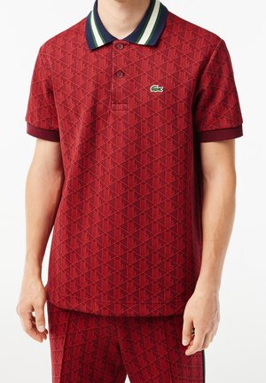 Rood poloshirt met patroon en een geribbelde marineblauwe kraag met groene en witte strepen. Korte mouwen met contrasterende manchetten. Lacoste-logo aanwezig.