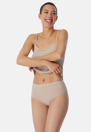 Smiling woman with short hair wearing beige tank top and high-waisted underwear, lifting her shirt slightly against a plain background.