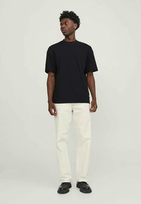 Homem de pé contra um fundo claro, vestindo uma camiseta preta, calças off-white e sapatos pretos, olhando para a sua esquerda.