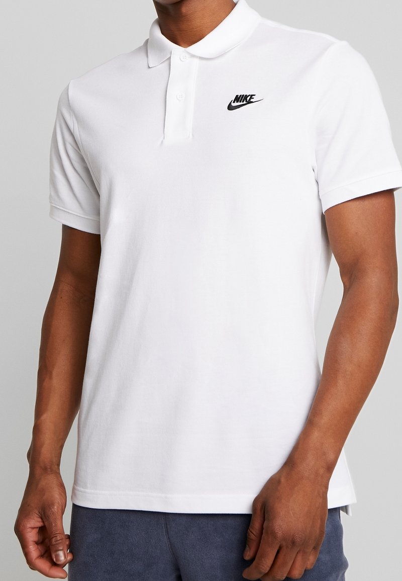 Homme portant un polo blanc Nike à manches courtes avec un logo Nike noir sur la poitrine gauche, associé à un pantalon foncé.