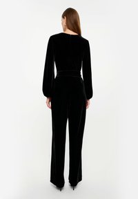 Czarny aksamitny kombinezon z długim rękawem, dopasowanym w pasie i szerokimi nogawkami. Posiada szew z tyłu oraz subtelny detal zamka na szyi.