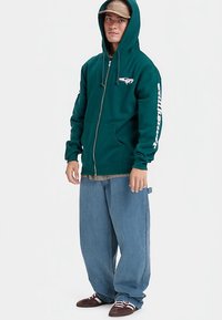 Jovem vestido com um hoodie verde com fecho, texto branco nas mangas, jeans azuis largos, sapatilhas castanhas e um chapéu bege com o capuz levantado.