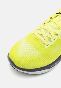 Kollane sportlik jalanõud, millel on hingav võrgust pealis, tekstuuriga detailid, kerged paelad ja must põhi, millel on nähtav logo ja pehme tehnoloogia.