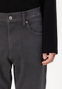 Grå jeans laget av denim med en glatt tekstur, med en frontlomme, metallknapp-lukking og sydd detaljer langs sømmene.