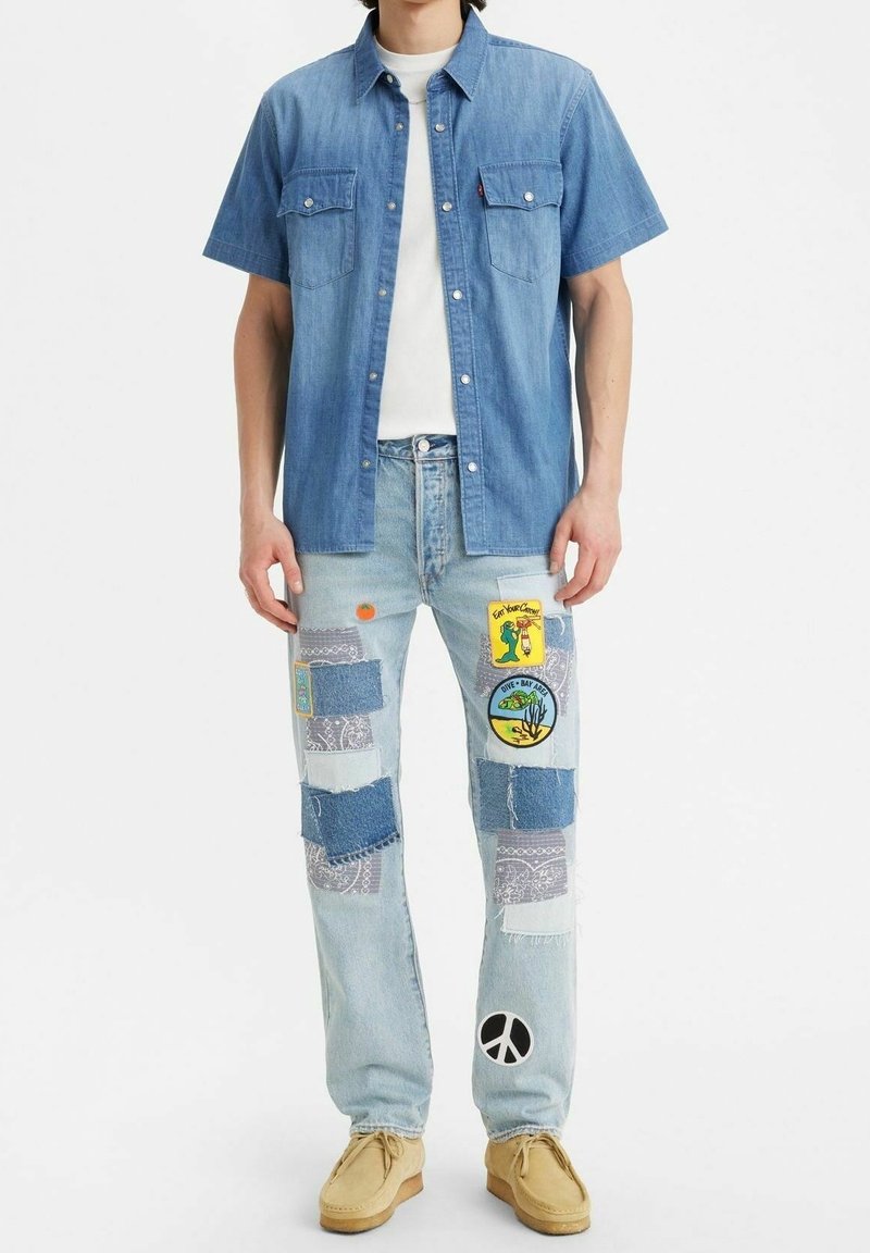 Camisa de manga corta de denim en azul claro con dos bolsillos en el pecho, combinada con pantalones de mezclilla de patchwork que presentan varias texturas, colores y parches impresos.