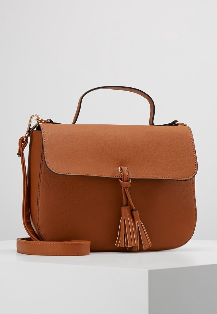 Hnědá kožená krosbody taška s klopou, detailem třásní, horním madlem a odnímatelným ramenním popruhem na bílé ploše a světle šedém pozadí.