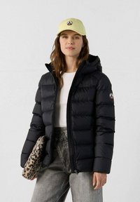 Kvinde iført en sort puffjakke, hvid skjorte, grå jeans og en lys gul kasket, som holder en clutchtaske med leopardprint mod en ensfarvet væg.